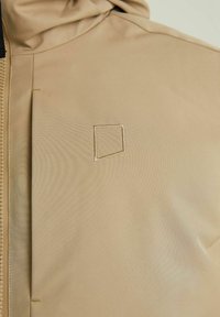 Beżowa kurtka z kapturem wykonana z gładkiej tkaniny, z kwadratowym haftem na lewej piersi i zamkiem błyskawicznym.