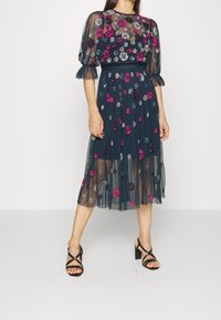 Mörkblå tylklänning med blommiga detaljer i rosa och silver. Har korta puffärmar och en midilång kjol. Svarta remsandaler.