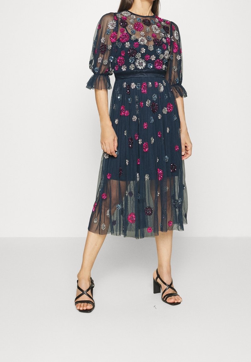 Mörkblå tylklänning med blommiga detaljer i rosa och silver. Har korta puffärmar och en midilång kjol. Svarta remsandaler.