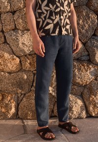 Dark denim pants with a straight leg fit, paired with a patterned short-sleeve shirt and black sandals, against a stone wall backdrop.