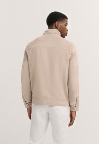 Beige jacket with a stand collar, long sleeves, and buttoned cuffs. Smooth texture with a simple design, worn over light-colored pants.