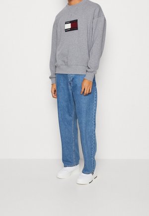 Szara bluza z kolorową naszywką z logo, zestawiona z luźnymi niebieskimi dżinsami i białymi trampkami. Codzienny look z miękkimi fakturami.