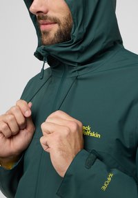 Veste imperméable verte avec capuche, dotée de cordons de serrage réglables, d'un logo jaune et de poignets Velcro pour un ajustement sécurisé et un confort optimal.