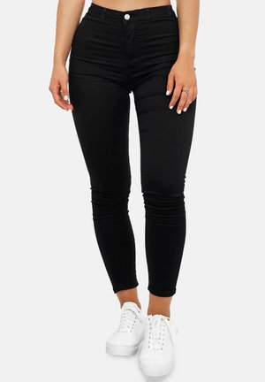 Vrouw in hoge black skinny jeans en witte sneakers, staand tegen een effen lichte achtergrond.