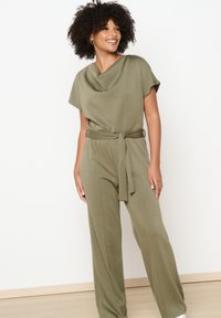 Zeleni kombinezon s kratkimi rokavi, narejen iz mehkega in raztegljivega blaga, z drapirano ovratnikom, pasom in širokimi hlačnicami.