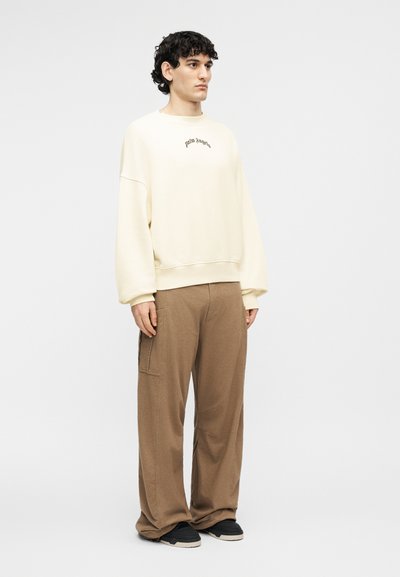 Νεαρός άνδρας φοράει υπερμεγέθη κρεμ φούτερ με το κείμενο "palm angels", καφέ παντελόνι φαρδιάς γραμμής και μαύρα αθλητικά παπούτσια σε λευκό φόντο.