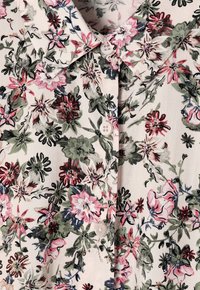 Blouse florale avec un fond crème clair, motif floral multicolore en rose, vert et noir, avec un col et des détails de boutons.