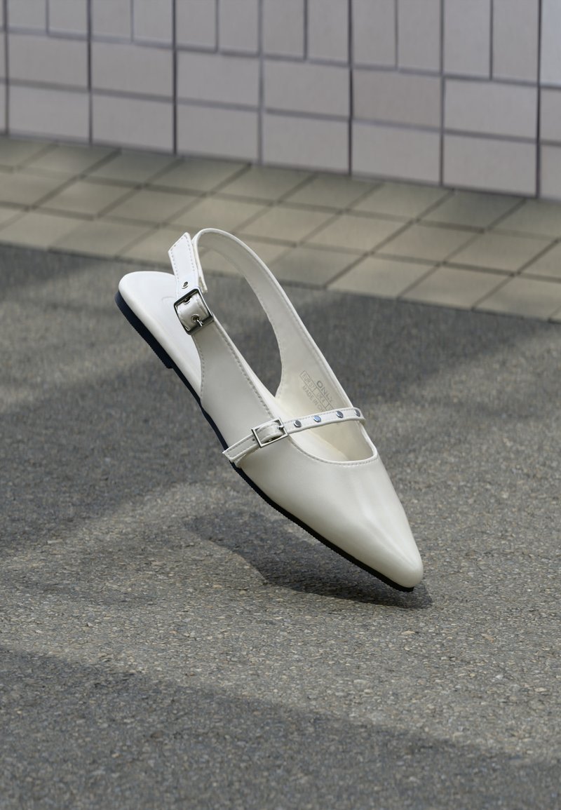 Λευκό μυτερό γυναικείο παπούτσι τύπου slingback με δύο ασημένιες αγκράφες, φωτογραφημένο σε εξωτερικό χώρο πάνω σε υφή εδάφους και πλακιδωτός τοίχος στο φόντο.