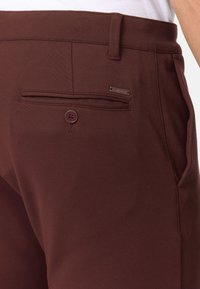 Bordeauxfarbene Hosen haben eine glatte Textur, einen taillierten Schnitt und eine hintere Tasche mit Knopf sowie einem kleinen Metalletikett der Marke.