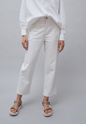 Persona che indossa jeans bianchi a gamba dritta, una camicia bianca con bottoni e sandali beige con punta aperta su sfondo grigio uniforme.