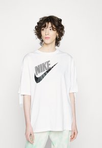 Camiseta blanca de gran tamaño hecha de algodón que presenta un gran logo negro de Nike con un diseño diagonal. Tiene mangas cortas con detalles de cordón.