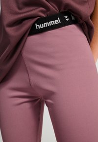 Paarse sportleggings met een soepele textuur, voorzien van een zwarte merkband aan de taille. Close-up toont de stikdetails en de strakke pasvorm.