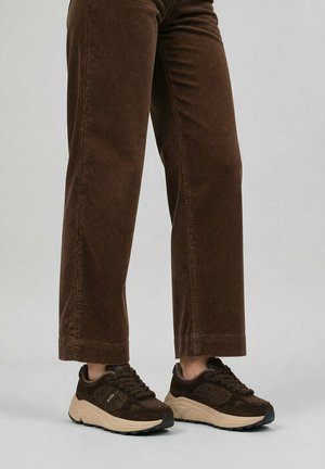 Persona que lleva pantalones de pana marrón y zapatillas marrón oscuro con suelas beige, de pie sobre un fondo liso.