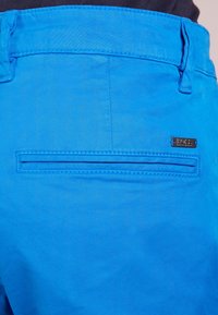 Blå shorts i lätt tyg med en bakficka med platt design och en liten metalltagg. Slät textur.