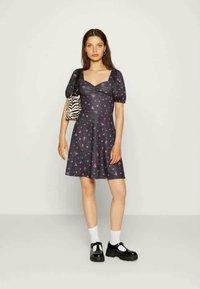 Vestido escuro, azul-marinho, com padrão floral, mangas bufantes, corpete ajustado e saia rodada. Modelo usa sapatos pretos e carrega uma bolsa riscada.