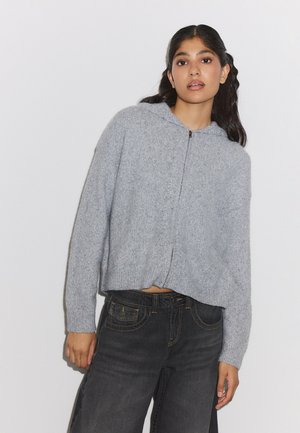 Jeune femme aux longs cheveux foncés porte un sweat à capuche zippé gris clair et un jean noir taille haute sur un fond blanc uni.