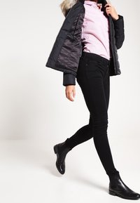 Czarna pikowana kurtka z kapturem wykończonym futrem, zestawiona z jasnoróżową koszulą w paski i czarnymi wąskimi dżinsami. Czarnie botki do kostek dopełniają strój.