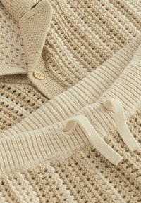 Cardigan beige tricoté avec un motif texturé, doté d'un bouton en bois et de poignets côtelés. Comprend un pantalon de jogger à cordon assorti.