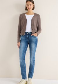 Mujer vistiendo un cárdigan marrón con botones sobre una camisa blanca, jeans azules y zapatillas de color claro, de pie contra una pared beige.