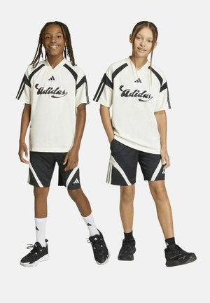 Dos niños con ropa deportiva Adidas blanca y negra a juego, de pie y sonriendo frente a un fondo sencillo.