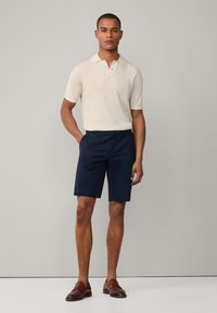 Béžová polokošeľa s krátkym rukávom a dvoma gombíkmi, skombinovaná s tmavomodrými šortkami. Model má obuté hnedé slip-on topánky. Jednoduchý a klasický dizajn.