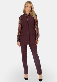 Bordowy kamizelka z dwoma przednimi kieszeniami noszona na wzorzystej bluzce, w połączeniu z dopasowanymi spodniami i czarnymi szpilkami. Gładka tekstura materiału.