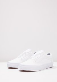 Une paire de baskets blanches basses propres avec des lacets et un léger détail sur les côtés, placée sur un fond blanc uni.