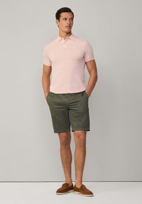 Svetle růžové polo tričko s krátkým rukávem, v kombinaci s olivově zelenými šortkami; nošeno s hnědými nazouvacími botami, jednoduchý design, hladká textura.
