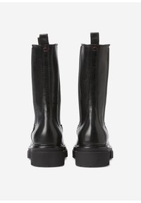 Ein Paar hohe schwarze Lederstiefel mit dicken Gummisohlen, von hinten gegen einen weißen Hintergrund betrachtet.