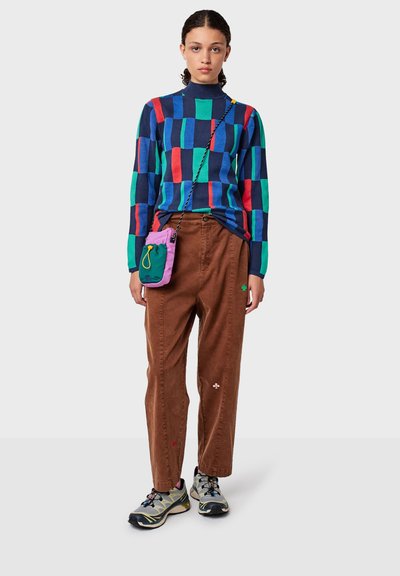 Femme portant un pull coloré à carreaux, un pantalon marron avec de petits symboles brodés, des baskets grises et un petit sac bandoulière violet et vert.