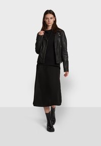 Veste en cuir noir de biker avec des accents matelassés sur les épaules, portée avec un haut noir et une jupe midi noire fluide. Des bottes de cheville noires complètent le look.