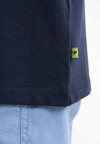 Camiseta de algodón en color azul marino con textura acanalada, que presenta una pequeña etiqueta amarilla con la letra "P". Detalles de las costuras visibles cerca del dobladillo.