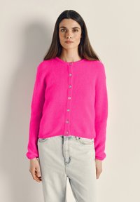 Moteris su ilgais rudais plaukais, vilkinti ryškiai rožinį sagotu megztinį ir šviesiai mėlynas džinsines kelnes, stovinti priešais paprastą šviesų foną.