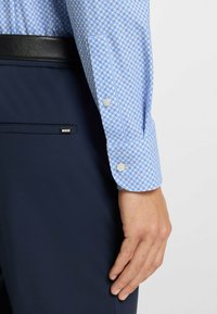 Vaaleansininen/malli vaatteissa pilkullinen kauluspaita, jossa on pienet pyöristetyt kuviot ja napitettavat hihat. Tummansiniset housut mustalla vyöllä ja logotägin kanssa.