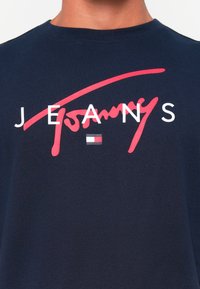 Námornícky modré bavlnené tričko s výrazným červeným kurzívou napísaným textom "Tommy" a bielym nápisom "JEANS", spolu s malým detailom logo pruhu.