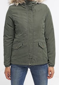 Zielona oliwkowa kurtka zapinana na zamek z kapturem wyłożonym sztucznym futrem, dwoma przednimi kieszeniami z klapkami na guziki oraz elastycznymi mankietami. Gładka faktura, casualowy styl.