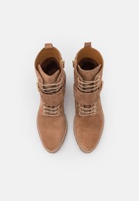 Stivaletti in suede marrone con punta arrotondata, lacci decorativi e dettagli in metallo dorato. Presentano una cintura e dettagli cuciti lungo la suola.