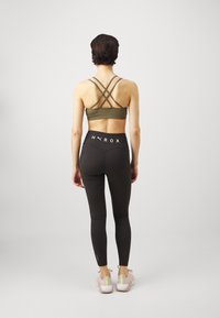 Reggiseno sportivo verde oliva con spalline incrociate, leggings neri a vita alta con testo bianco "H Y R O X" e sneakers rosa chiaro.