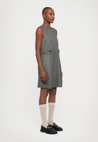 Ärmelloses graues karierter Kleid mit ausgestellter Silhouette, zwei Seitenknöpfen und glatter Textur. Kombiniert mit schwarzen Slippern und kniehohen Socken.