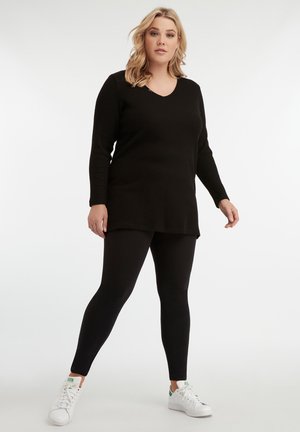 Zwarte gebreide tuniek met een V-hals en lange mouwen, gecombineerd met zwarte leggings en witte sneakers; soepele textuur en ontspannen pasvorm.