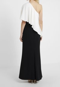 Kobieta w czarnej sukni do ziemi z białym asymetrycznym rękawem na jednym ramieniu, stojąca na tle jednolitego białego tła.