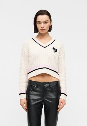 Ung kvinde stående iført en cremefarvet kort kabelstrikket sweater med sorte og pink striber og sorte læderbukser mod en ensfarvet baggrund.