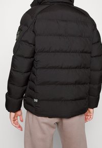 Černá péřová bunda s vodorovným prošíváním, se zipem a štítkem na dolní levé zadní části. Vybavena vysokým límcem a elastickými manžetami.