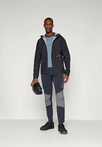 Μαύρο αδιάβροχο μπουφάν με γκρι λεπτομέρειες, συνδυασμένο με μπλε πουκάμισο και σκούρα παντελόνια με γκρι πάνελ. Περιλαμβάνονται μαύρα αθλητικά παπούτσια και κράνος ποδηλάτου.