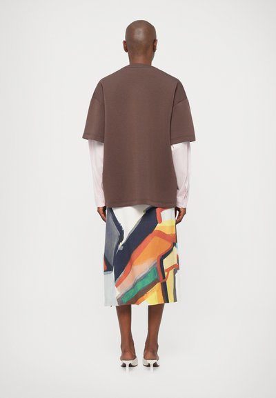 Brązowa, oversize'owa koszulka nałożona na długą, wielokolorową spódnicę o abstrakcyjnym wzorze, uszytą z lekkiego materiału, z białymi sandałkami na cienkich paskach.