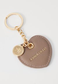 Llavero en forma de corazón hecho de faux cuero texturizado en taupe, con herrajes en tono dorado y una etiqueta de logo unida a una cadena.
