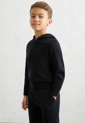 Čierna mikina s kapucňou s predným vačkom na ruky (kangaroo vreckom), dlhými rukávmi a voľným strihom. Materiál pôsobí mäkko a textúrovane, doplnený o zodpovedajúce nohavice.