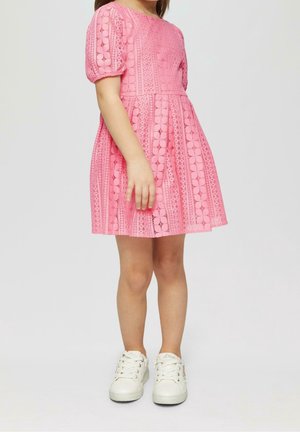 Enfant portant une robe en dentelle rose à manches courtes et des baskets blanches, debout devant un fond clair uni.