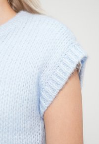 Ανοιχτό μπλε πλεκτό πουλόβερ με κοντά, γυριστά μανίκια. Το ύφασμα είναι μαλακό και αφράτο, με υφασμένο μοτίβο και ριγωτές λεπτομέρειες.