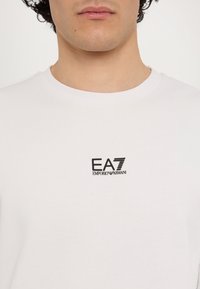 Hvid bomulds t-shirt med rund hals og korte ærmer. Sort EA7 Emporio Armani logo på brystet. Glat tekstur, enkelt design.